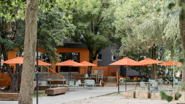 An outdoor seating area with orange umbrellas, tables, and chairs, surrounded by trees and a building with a modern design. - Combo Bologna - Hostel (Bologna)
