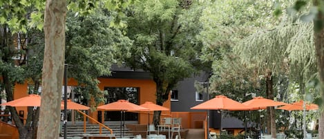Un área de estar al aire libre con sombrillas naranjas, mesas y sillas, rodeada de árboles y un edificio de diseño moderno.