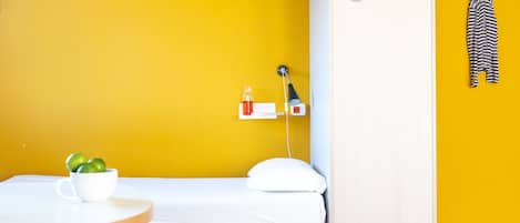 Una habitación con una cama, una mesita con una taza y limones, un estante con una lámpara, y una prenda a rayas colgada en la pared.