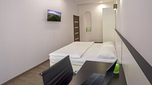 Hotellihuone, jossa on sänky, työpöytä kannettavalla tietokoneella, tuoli, seinälle asennettu televisio ja vihreä maljakko yöpöydällä.