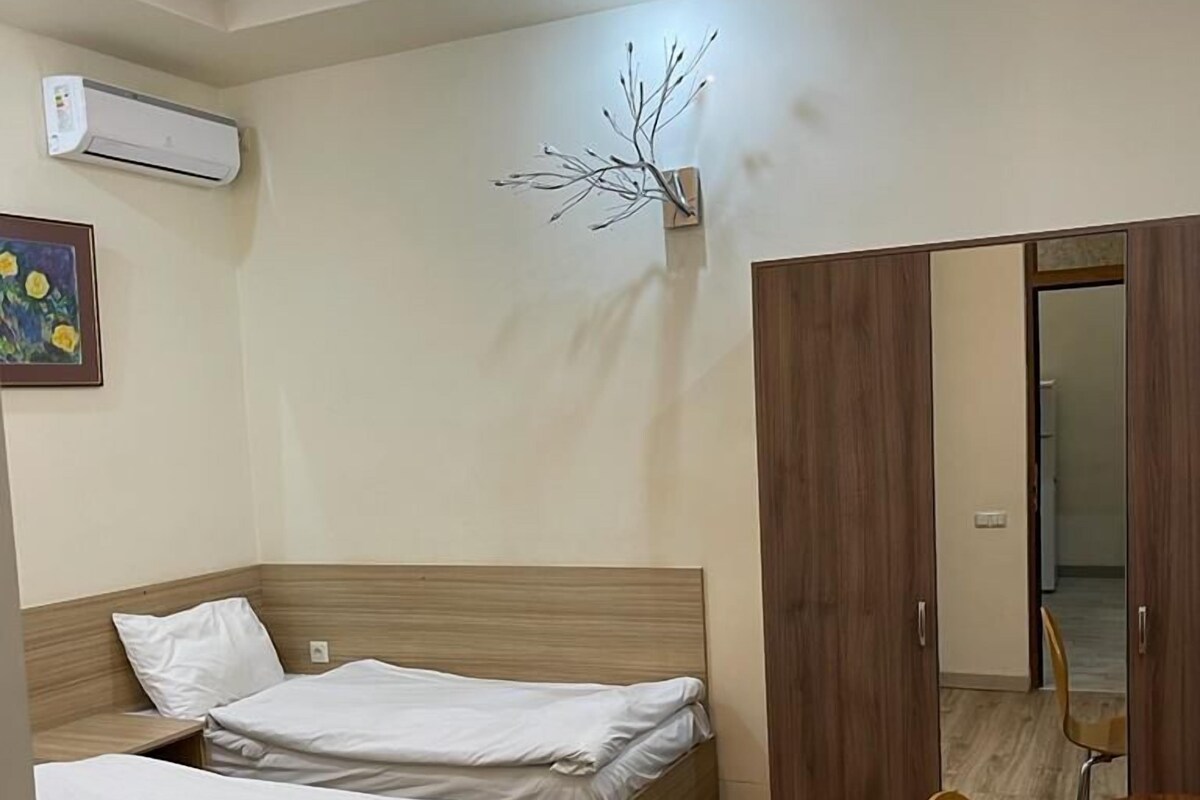 Ein Hotelzimmer mit zwei hölzernen Betten, einem kleinen Tisch mit Stühlen, einer wandmontierten Klimaanlage und einem dekorativen Zweig an der Wand.