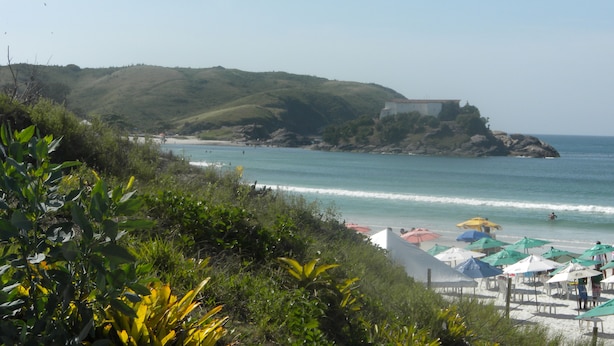 A beach with umbrellas and a white building on a hill. - Casa Nostra Hospedagem Residencial (Cabo Frio)
