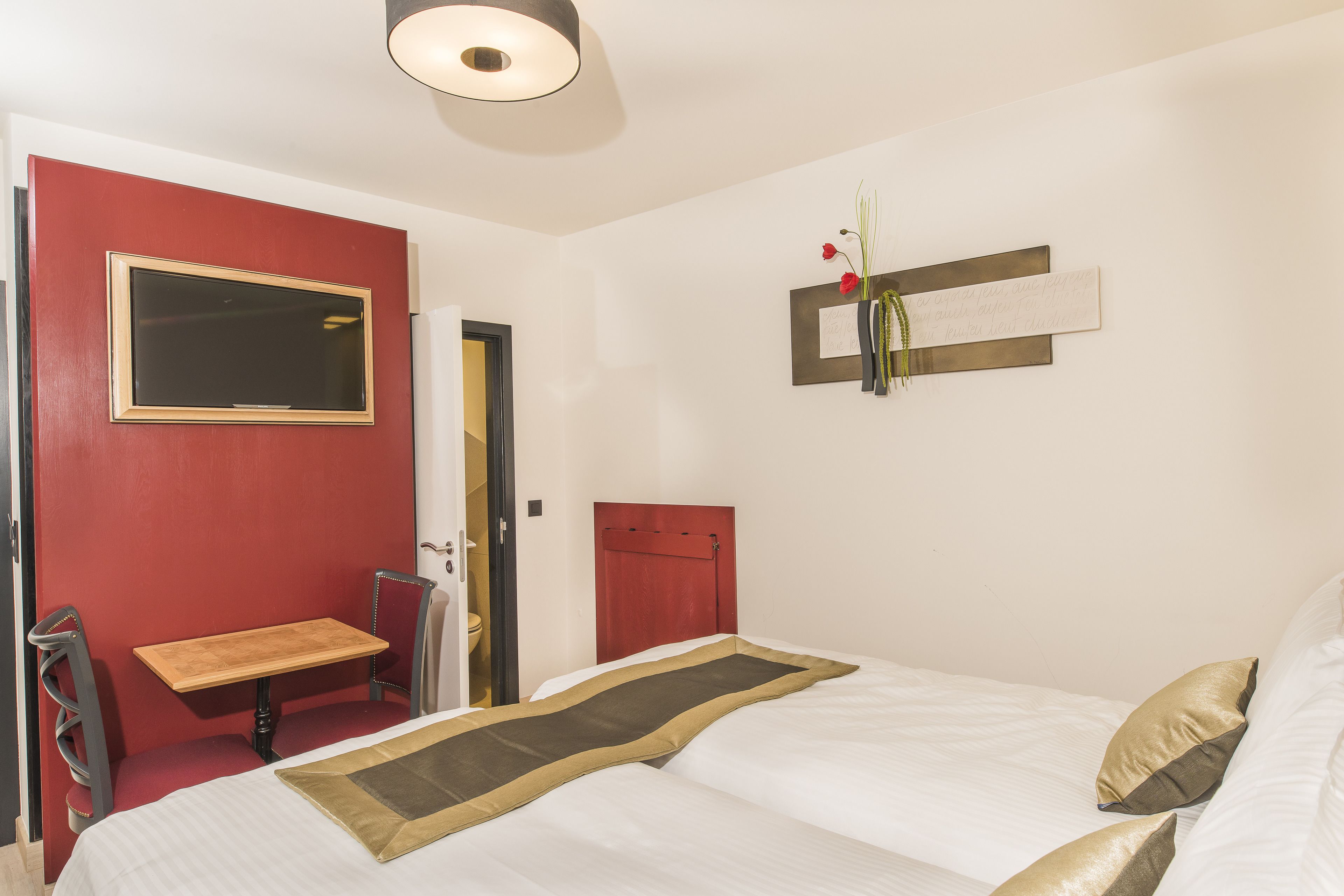 A hotel room with a bed, a small table, a chair, a mirror, and a red wall.