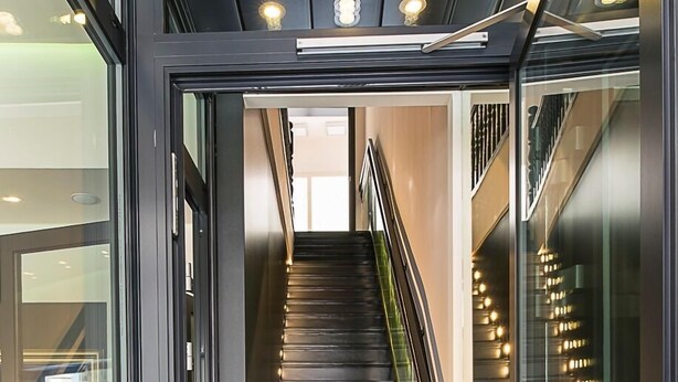 Una entrada de hotel moderna con puerta de cristal, una escalera que conduce a los pisos superiores y un felpudo con el nombre del hotel.