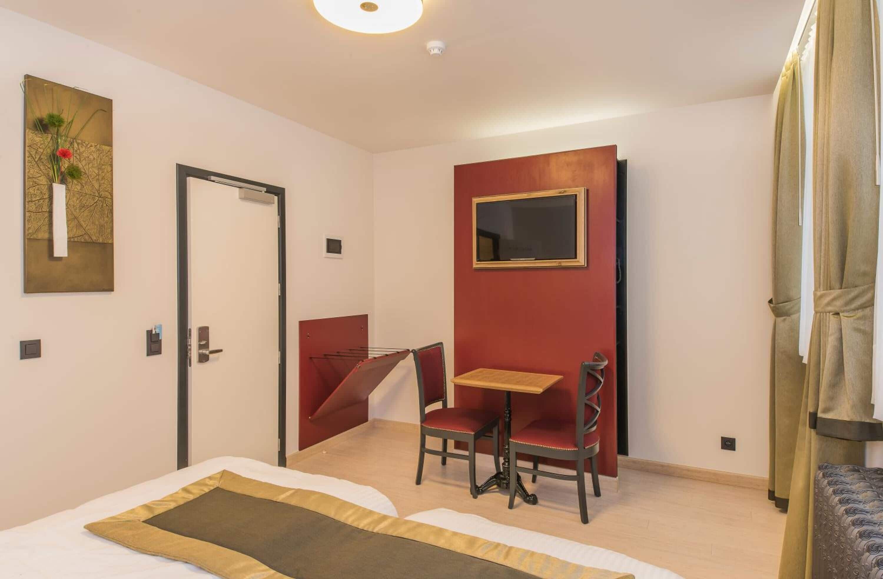 A hotel room with a bed, a small table, a chair, a mirror, and a red accent wall.