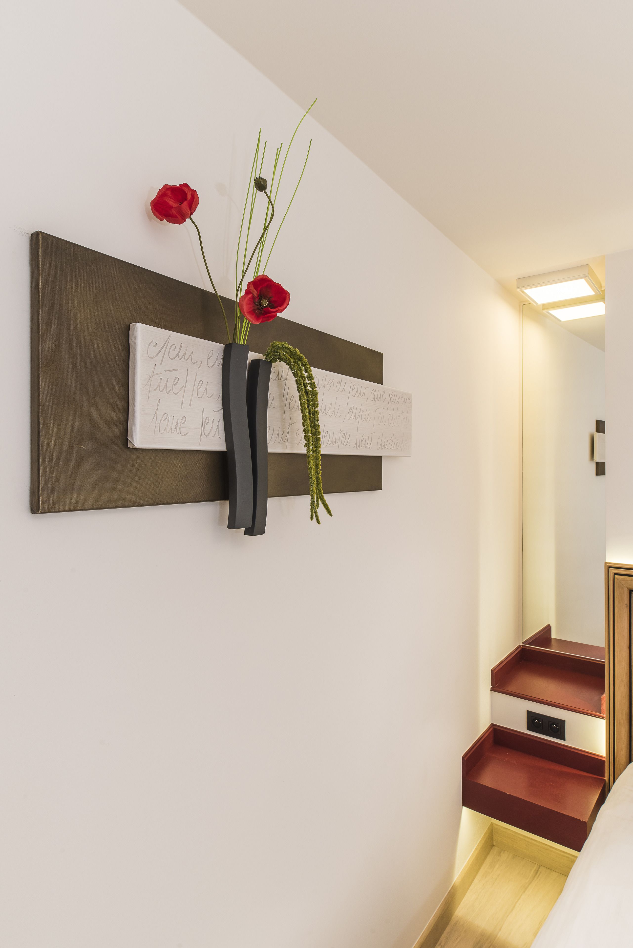 A modern hotel room with a minimalist design, featuring a wall-mounted artwork with text and a vase with red flowers.