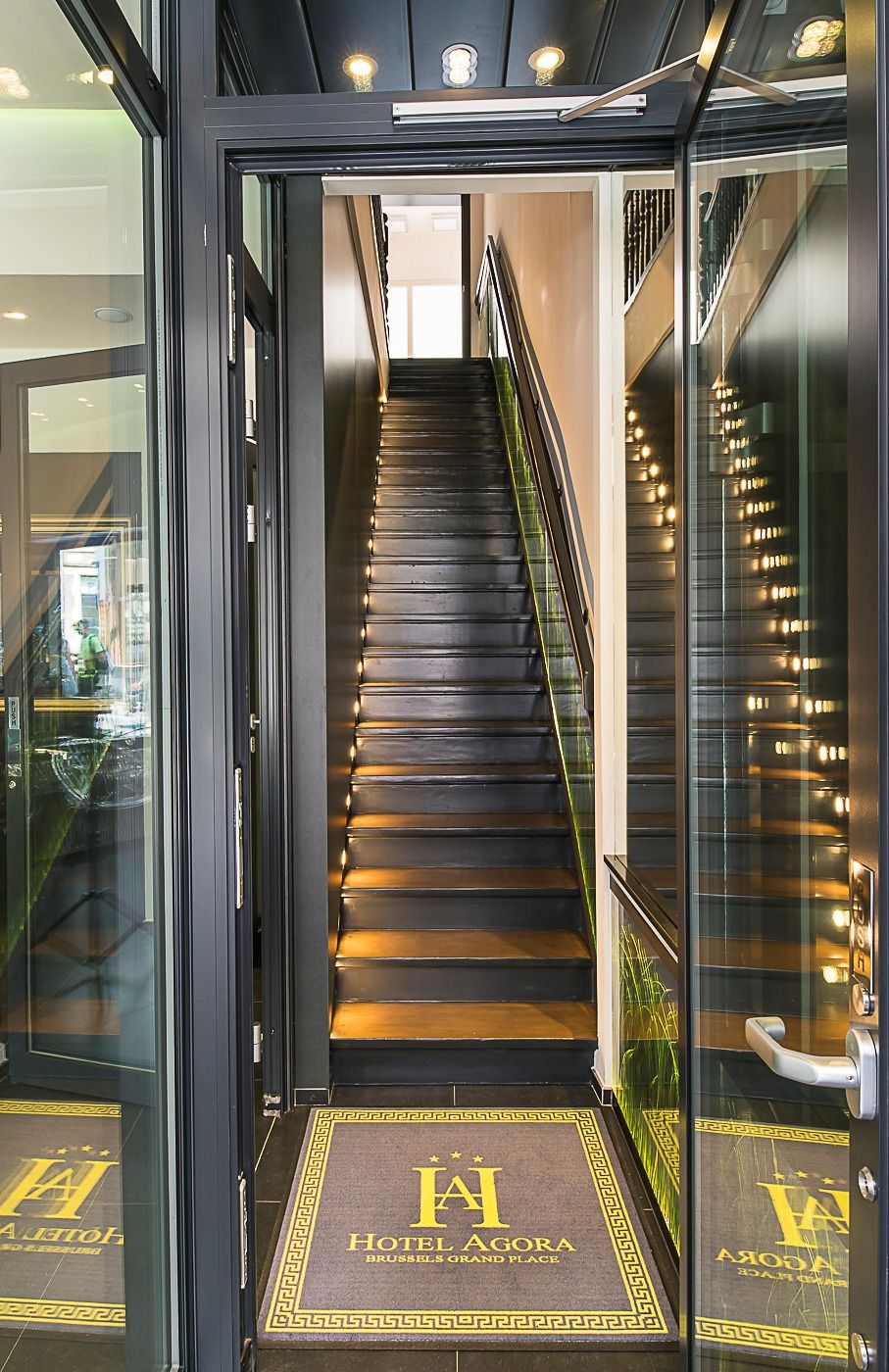 Entrada d'un hotel modern amb una porta de vidre, una escala que mena al pis superior i un felpú amb el nom de l'hotel.