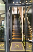 Una entrada de hotel moderna con puerta de cristal, una escalera que conduce a los pisos superiores y un felpudo con el nombre del hotel.