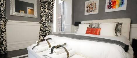 A modern hotel room with a large bed, a bedside table with towels, a mirror, and artwork on the walls.