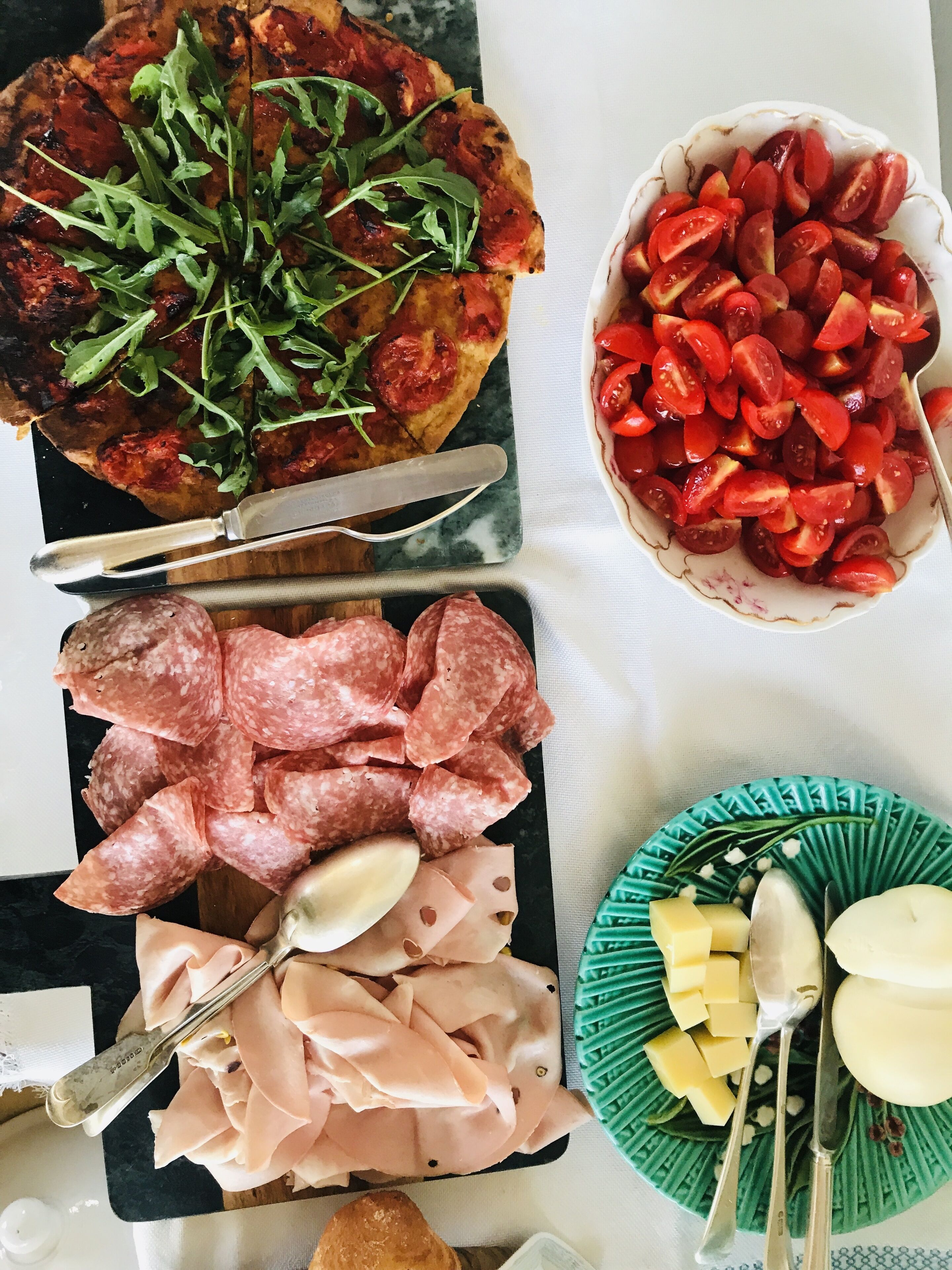 A variety of food items including pizza, sliced sausages, salami, cherry tomatoes, and cheese on a table.