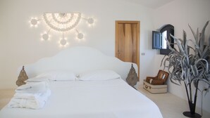 A neatly made bed with white linens, a wooden door, a plant, and a wall-mounted light fixture.