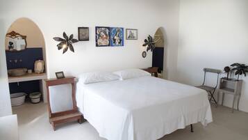 Une chambre à coucher avec un grand lit, une petite table, une chaise et une étagère ornée d’objets décoratifs.