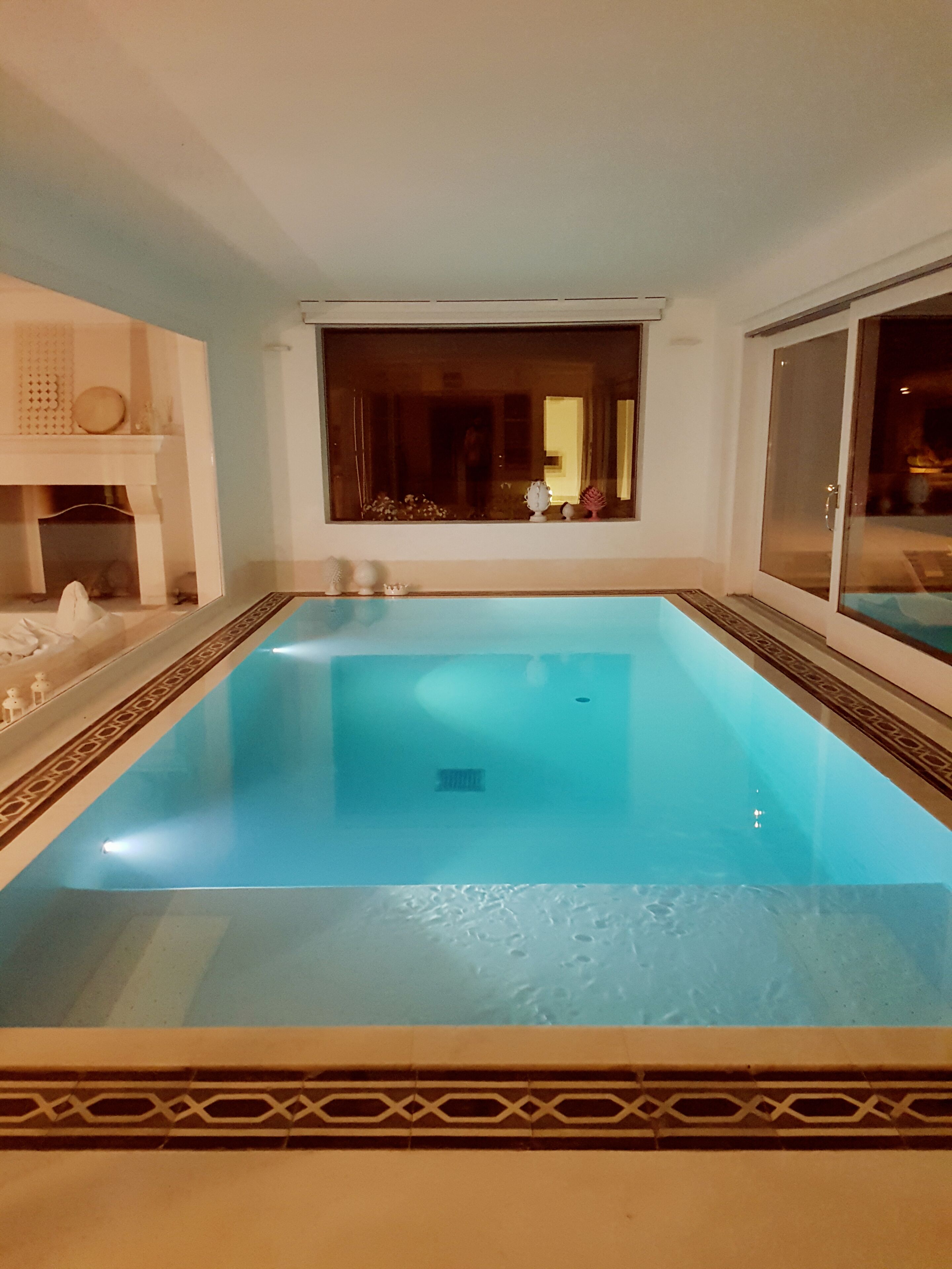 Una piscina interior rectangular con superficie de agua azul cristalina, rodeada por un borde decorativo de azulejos.