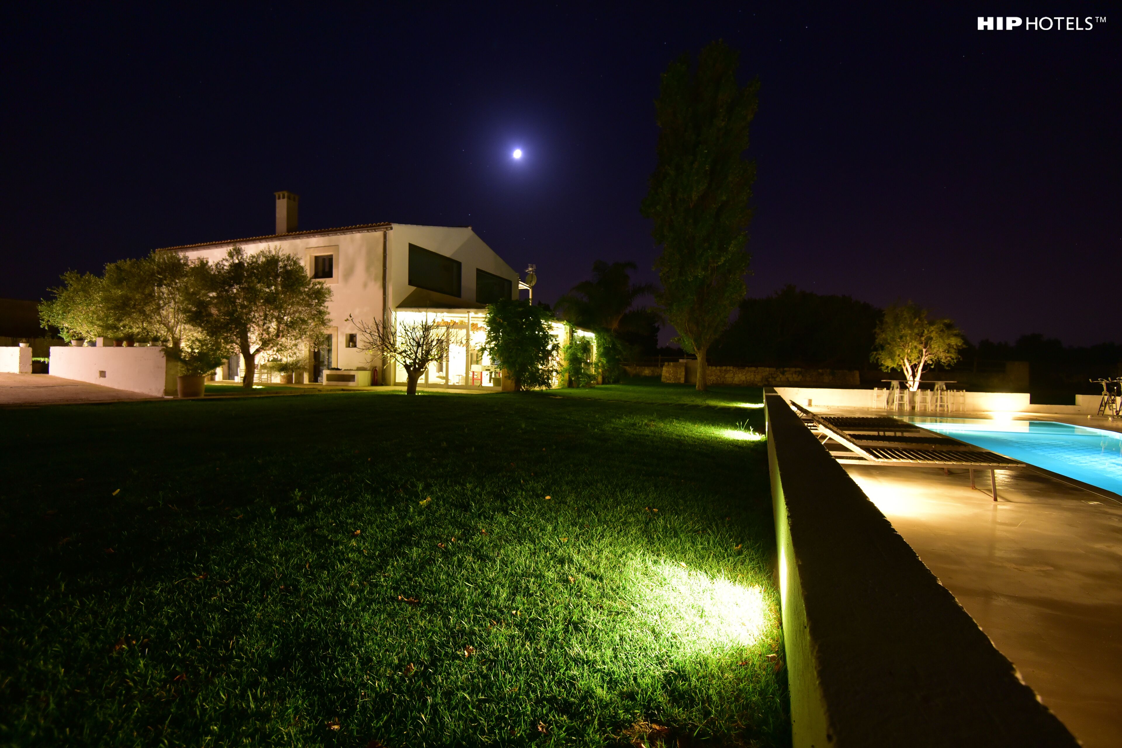 A well-lit outdoor area with a swimming pool, a lawn, and a building with a moon in the background.