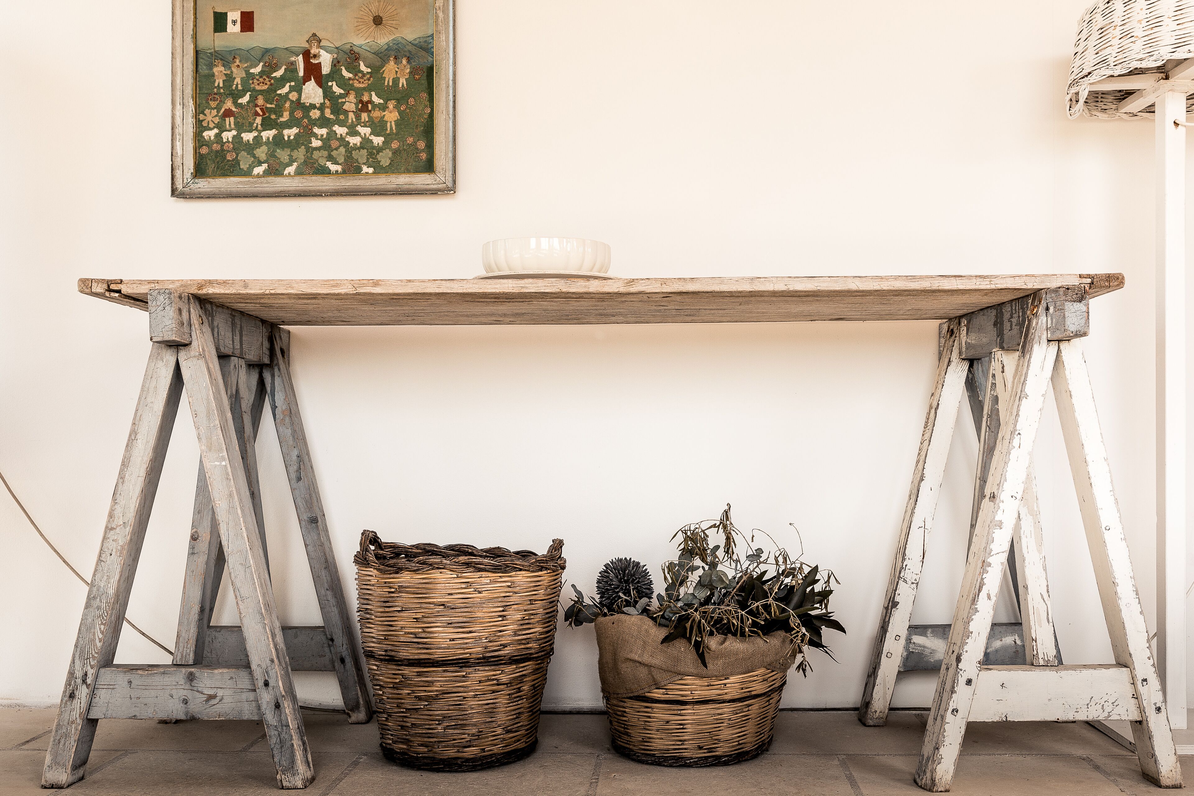 A rustic wooden table with a white bowl on top, flanked by two wicker baskets, one filled with plants and the other empty, against a plain wall.
