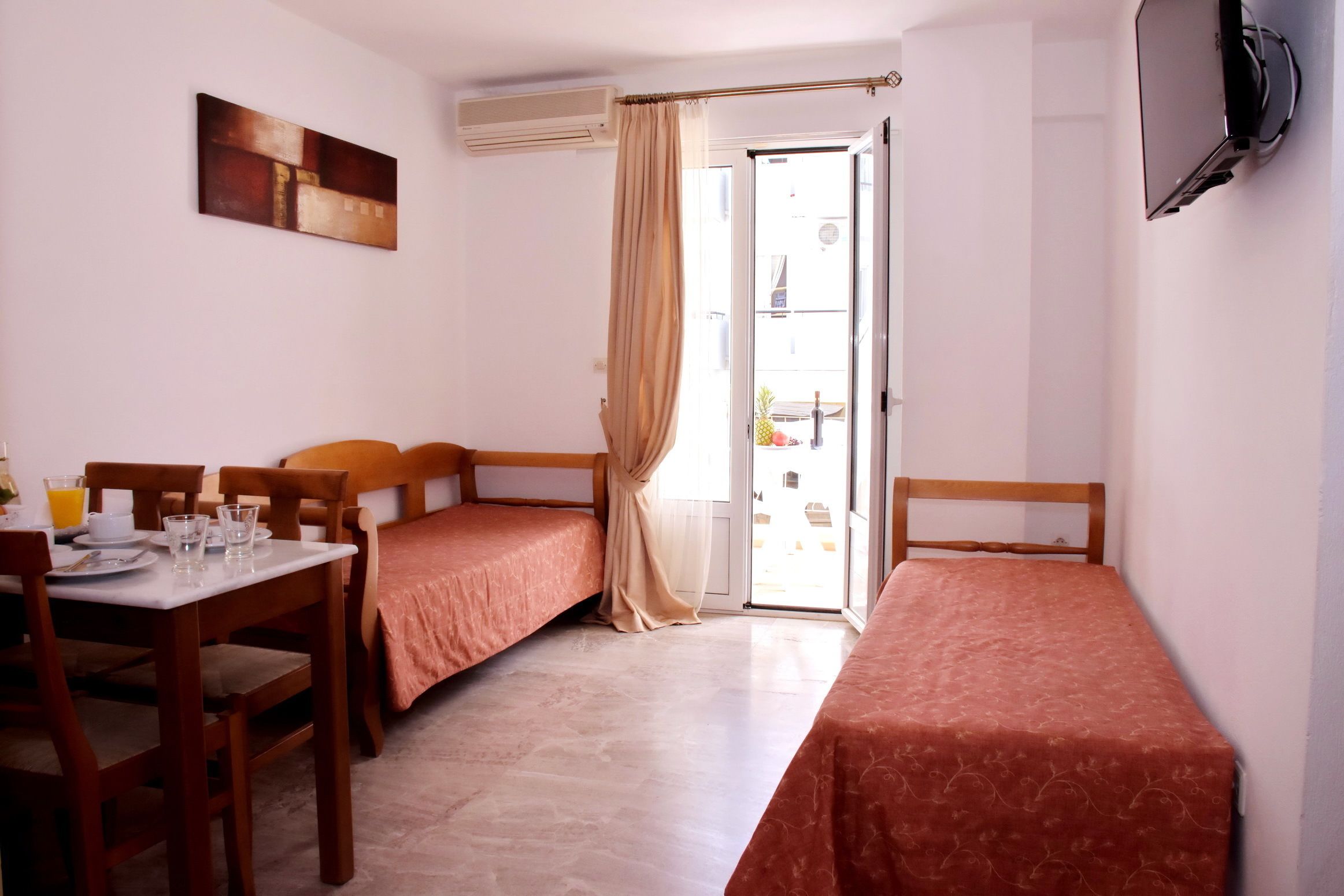 A room with two single beds, a dining table with chairs, a TV mounted on the wall, and a sliding glass door leading to an outdoor area.
