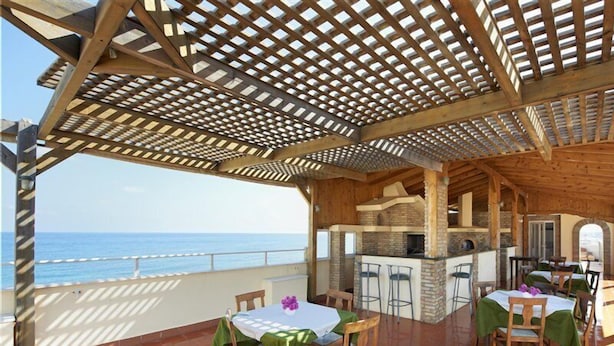 An outdoor patio with wooden tables and chairs, a brick oven, and a view of the sea. - Irini Apartments (Hersonissos)