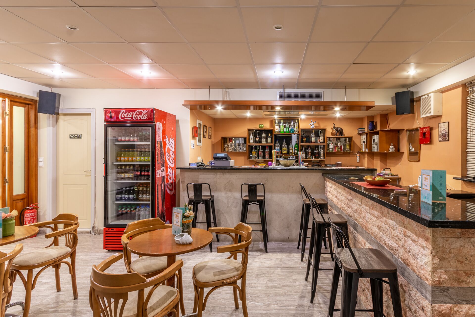 A bar area with a Coca-Cola refrigerator, wooden chairs, a marble counter, and shelves with bottles.