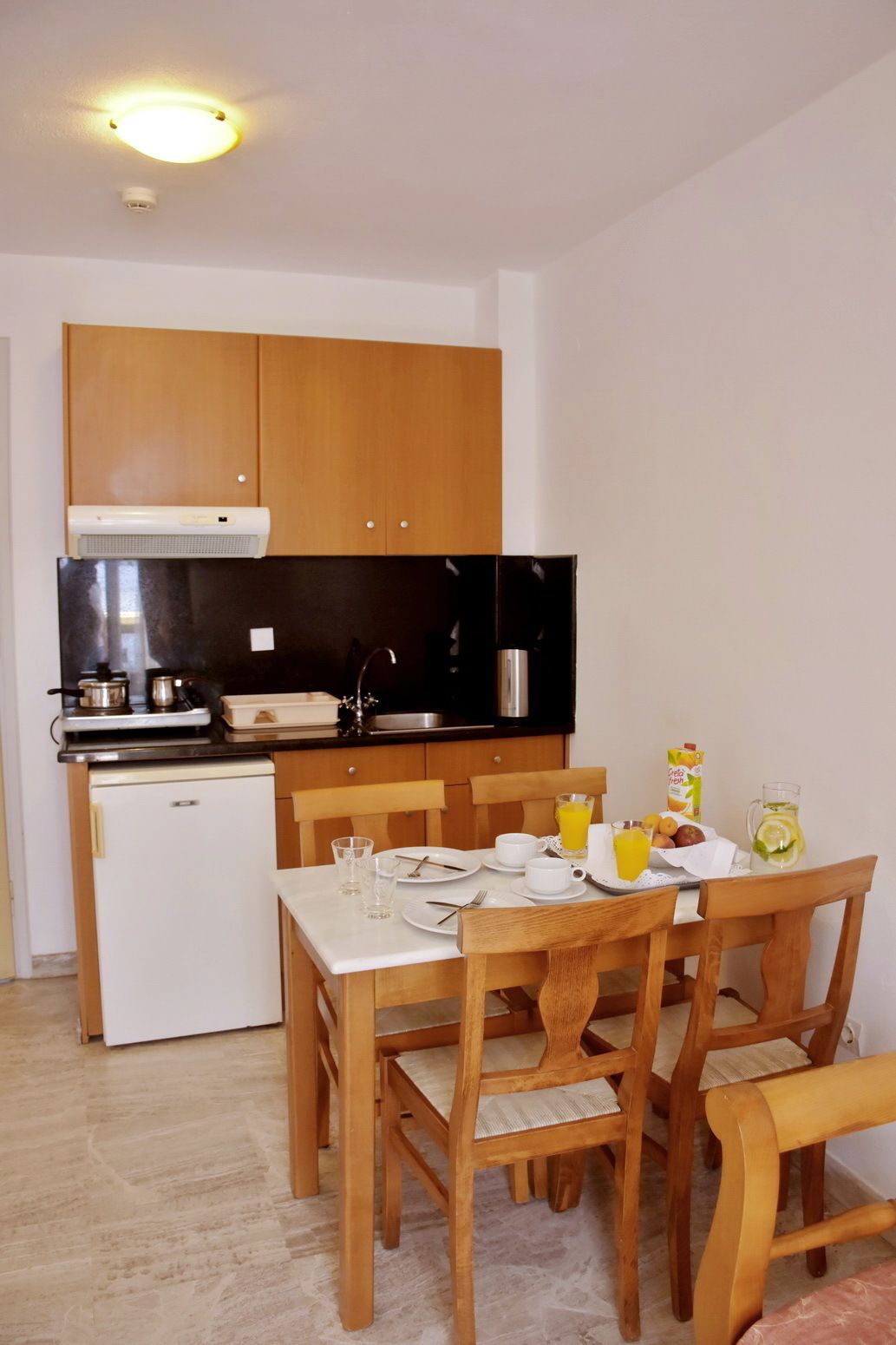 A compact kitchen with wooden cabinets, a black countertop, and a small refrigerator. A dining table with chairs is set for breakfast.