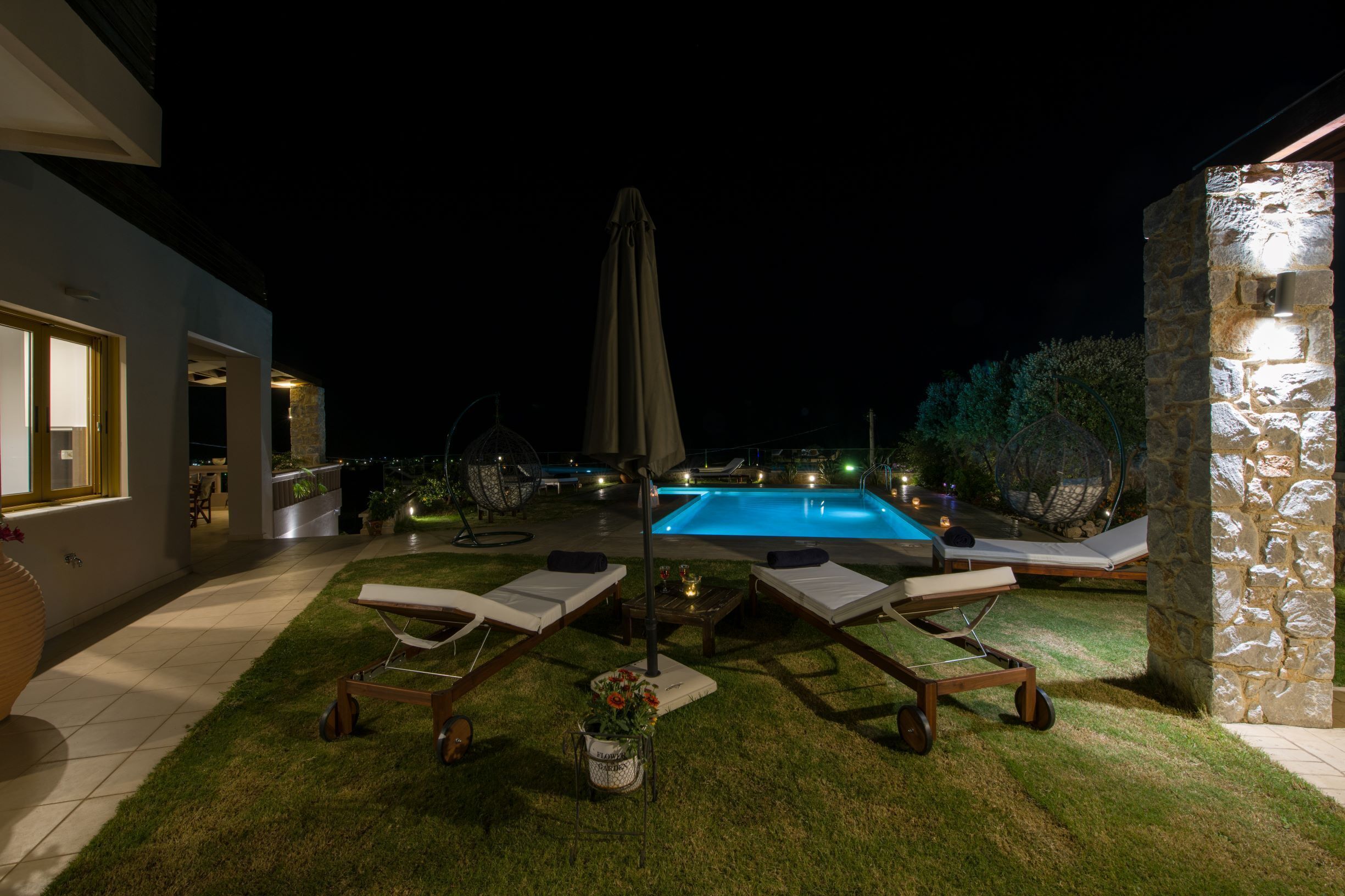 A nighttime outdoor pool area with lounge chairs, a table with a lamp, and a stone wall.