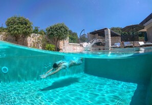 A swimmer in a clear pool with a stone wall and lounge chairs in the background. - Lady Dafni (Platanias)