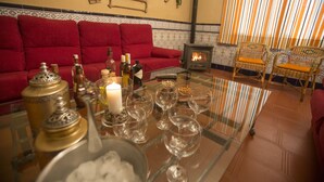 Una sala de estar acogedora con un sofá rojo, una mesa de centro de vidrio con copas y botellas de vino, una chimenea pequeña y sillones de mimbre.