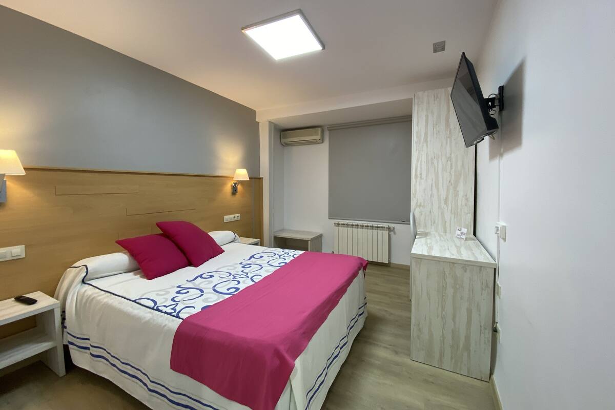 Een hotelkamer met een bed, nachtkastjes, een aan de muur bevestigde televisie en een raam met zonwering.
