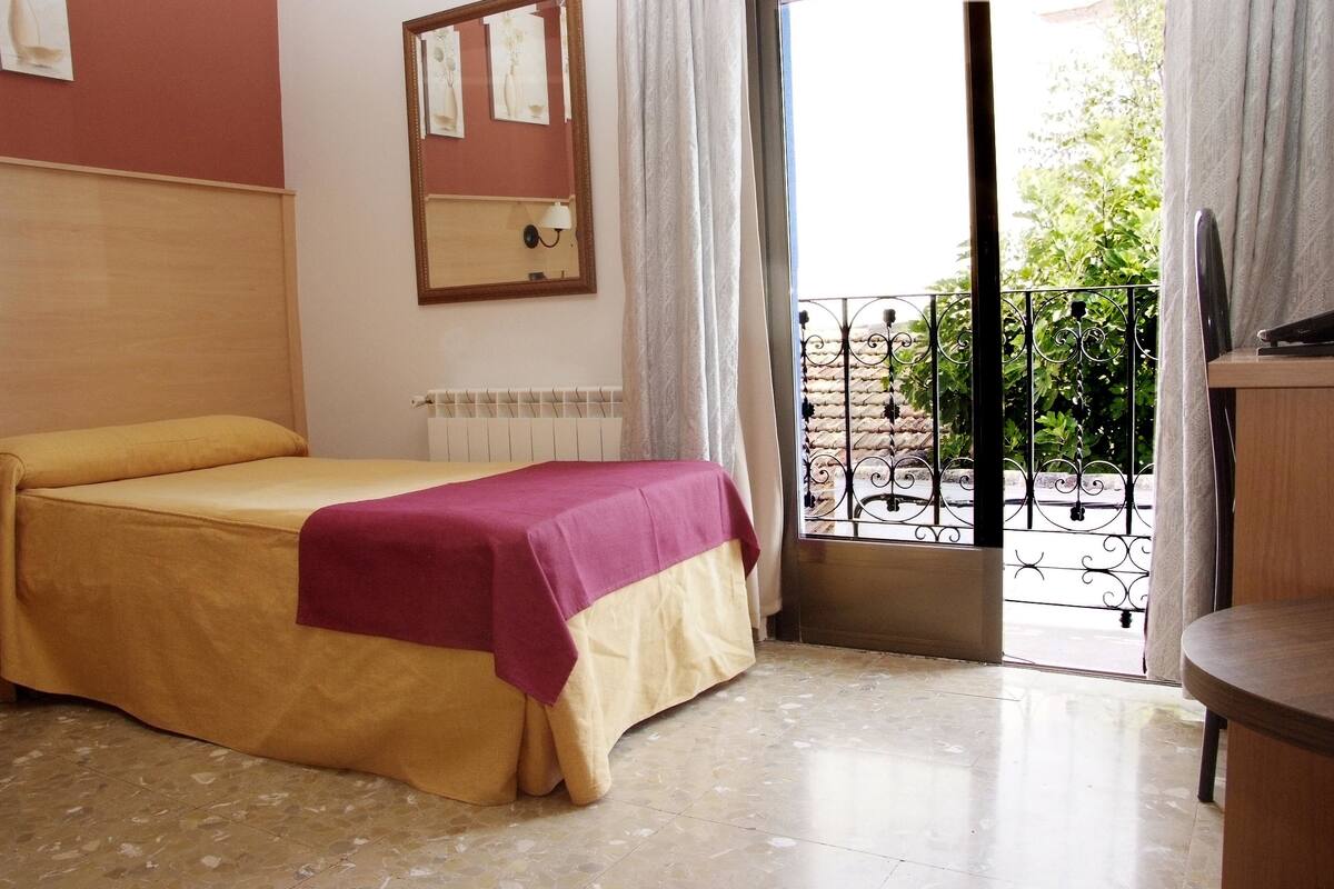 Hotelkamer met een bed, een klein tafeltje, een radiator, een raam met gordijnen en uitzicht op het balkon.