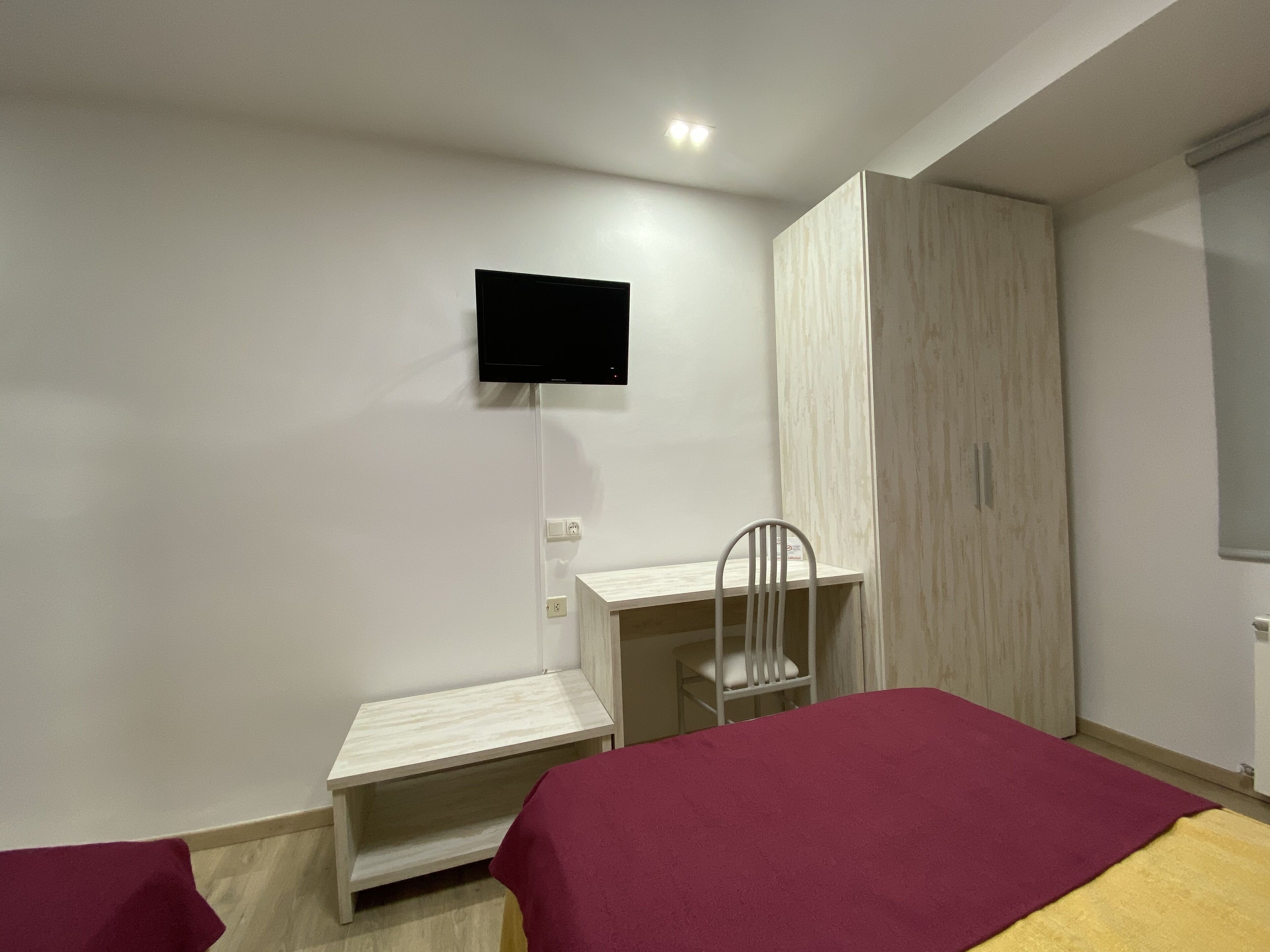 Een slaapkamer met een bed, een bureau, een stoel, een kledingkast en een aan de muur bevestigde televisie.