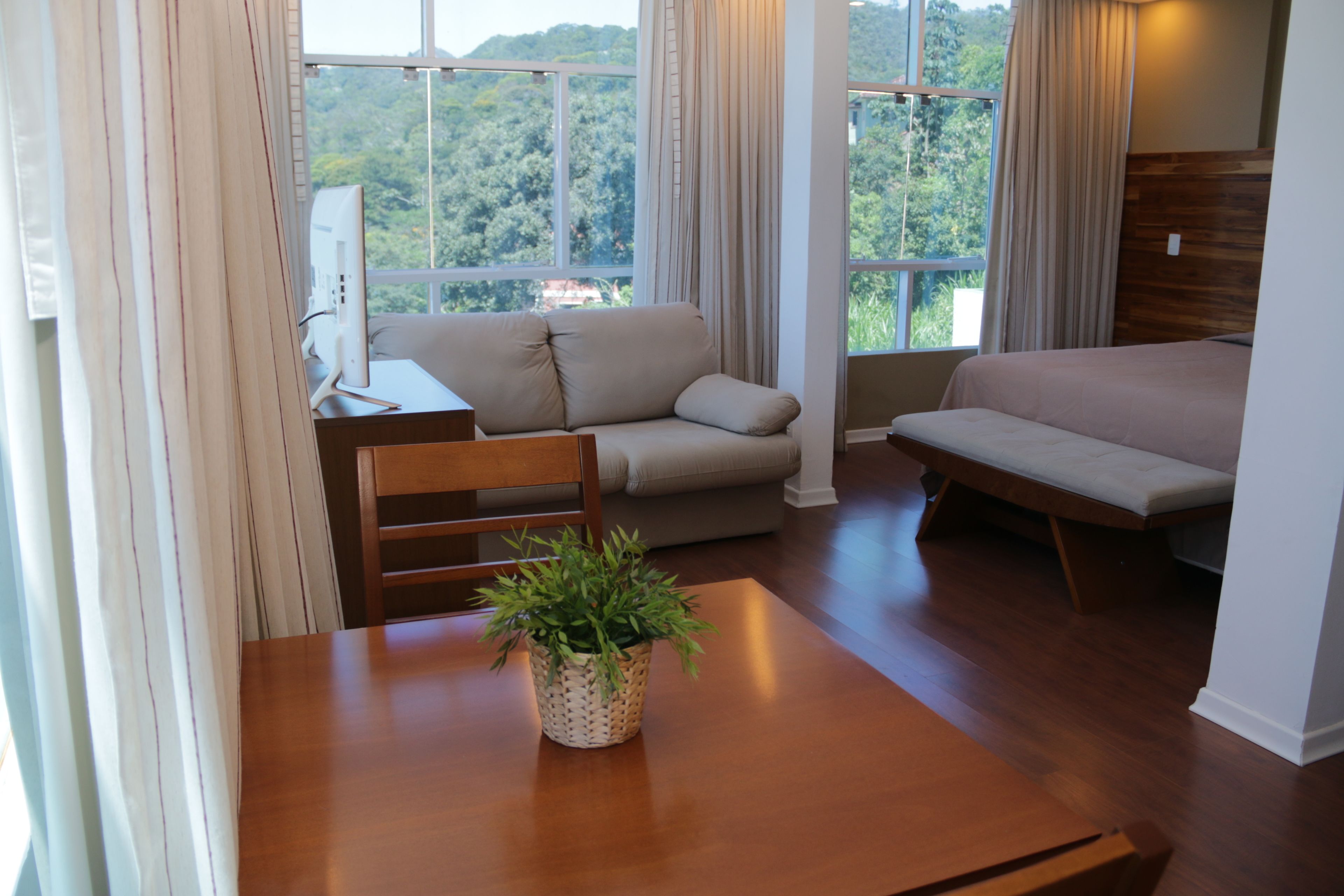 A room with a wooden table, a sofa, a bed, and a window with a view of greenery.