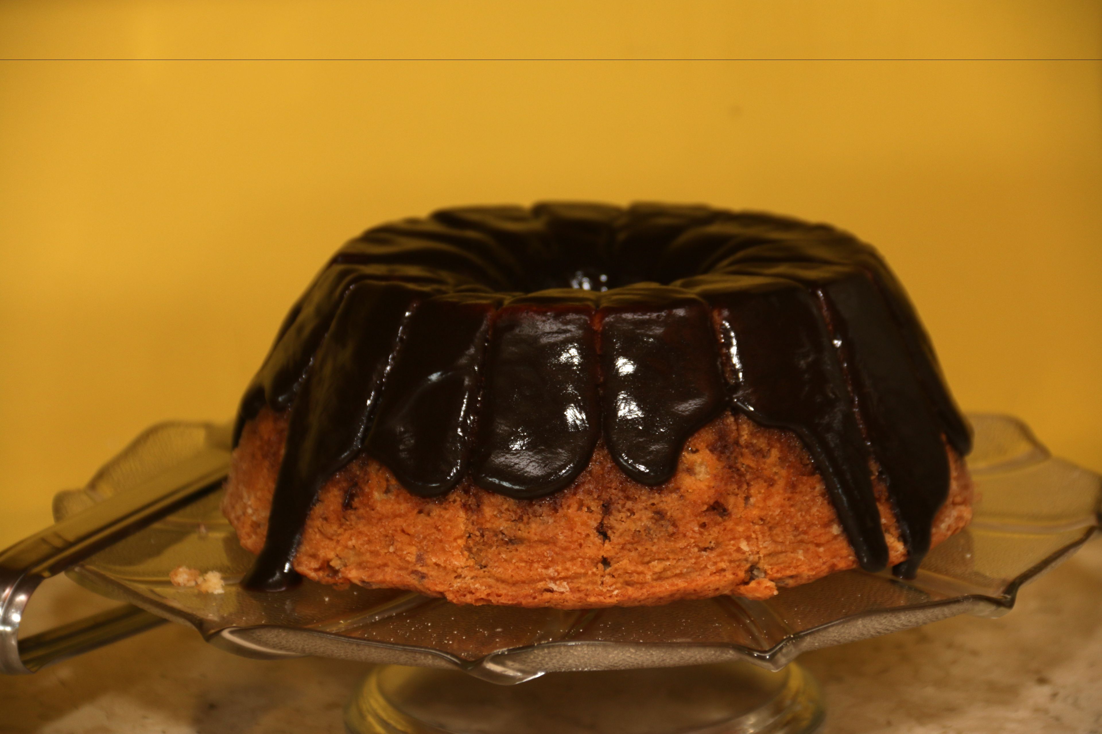 A bundt cake with chocolate glaze on a clear glass cake stand.
