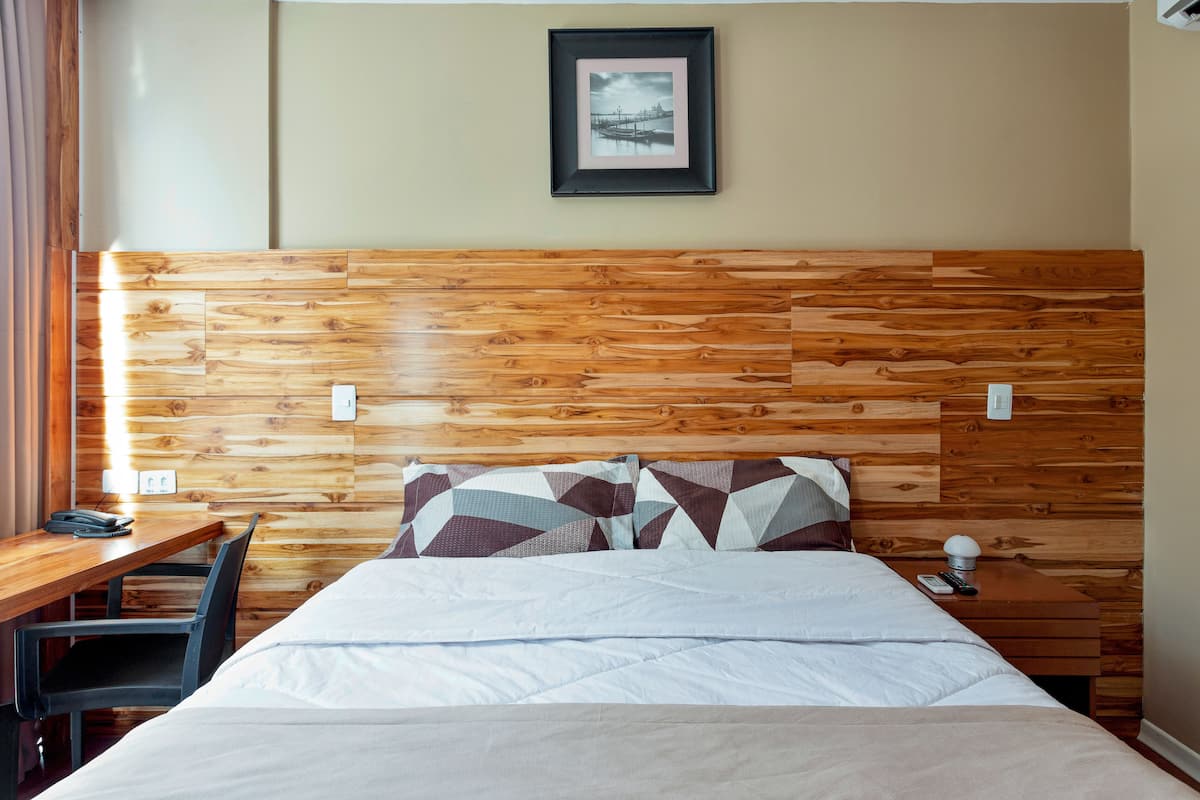 Habitación de hotel con cabecera de madera, cama con almohadas estampadas, mesita de noche con lámpara y un cuadro en la pared.