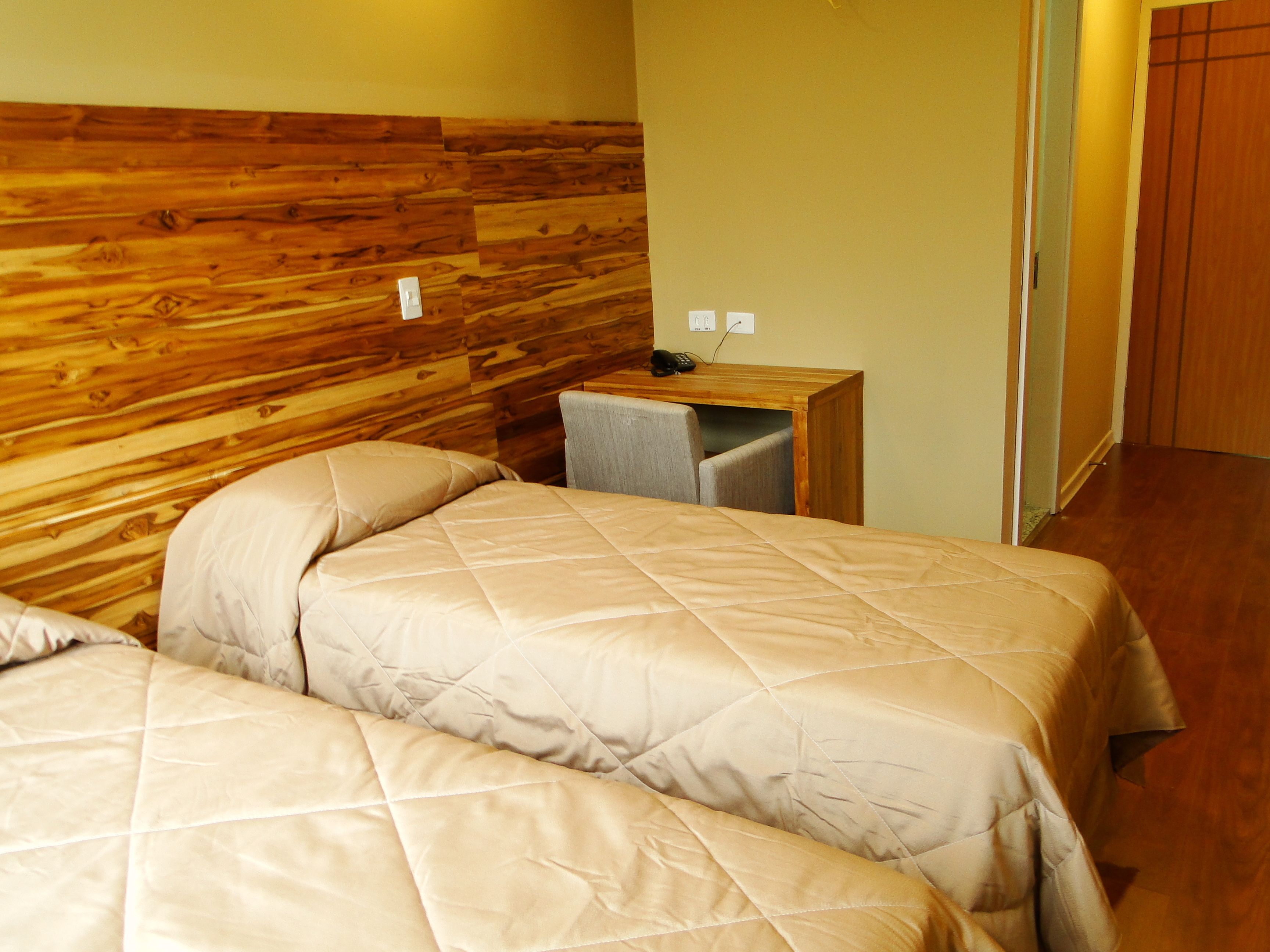 Habitación de hotel con dos camas, un cabecero de madera, un escritorio pequeño con una silla y una puerta que da a otra habitación.