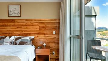 Quarto de hotel com cabeceira de madeira, cama com almofadas estampadas, mesa de cabeceira com abajur e varanda com cadeira de vime.