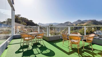 Una terraza en la azotea con sillas y mesas de mimbre, con vistas a montañas lejanas y una ciudad.