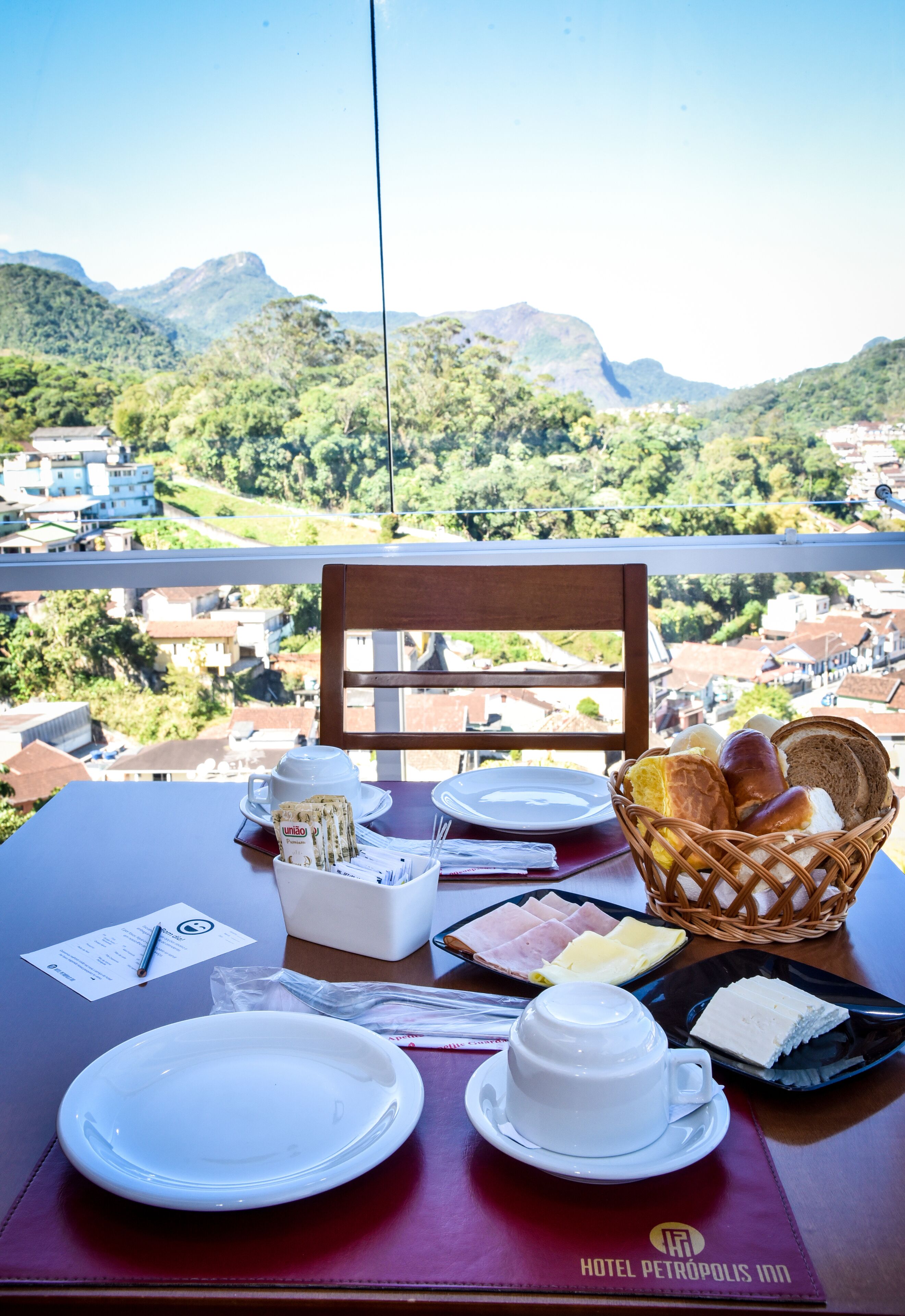 A table set with plates, a cup, a basket of bread, and a tray of food, overlooking a mountainous landscape.