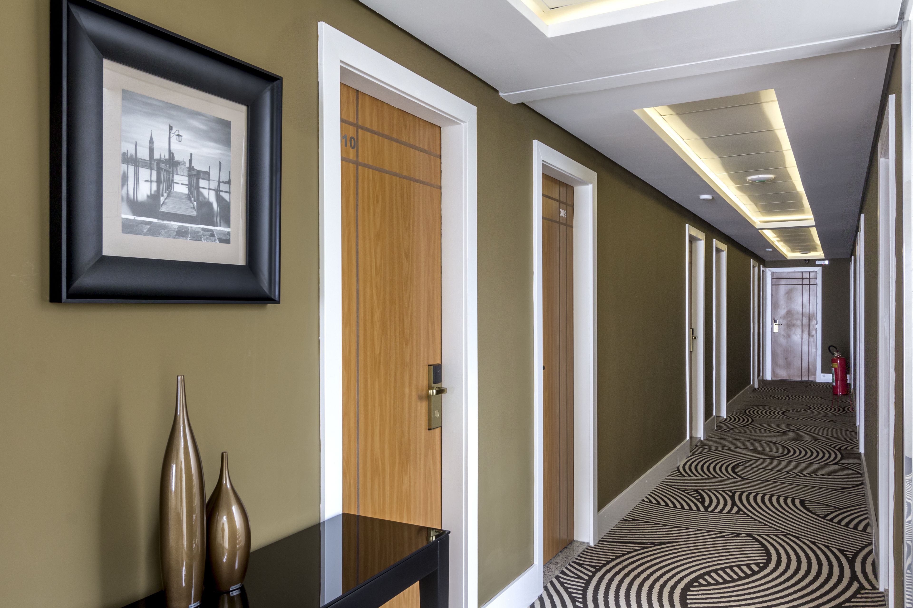 A hotel hallway with framed artwork, wooden doors, and patterned carpet.