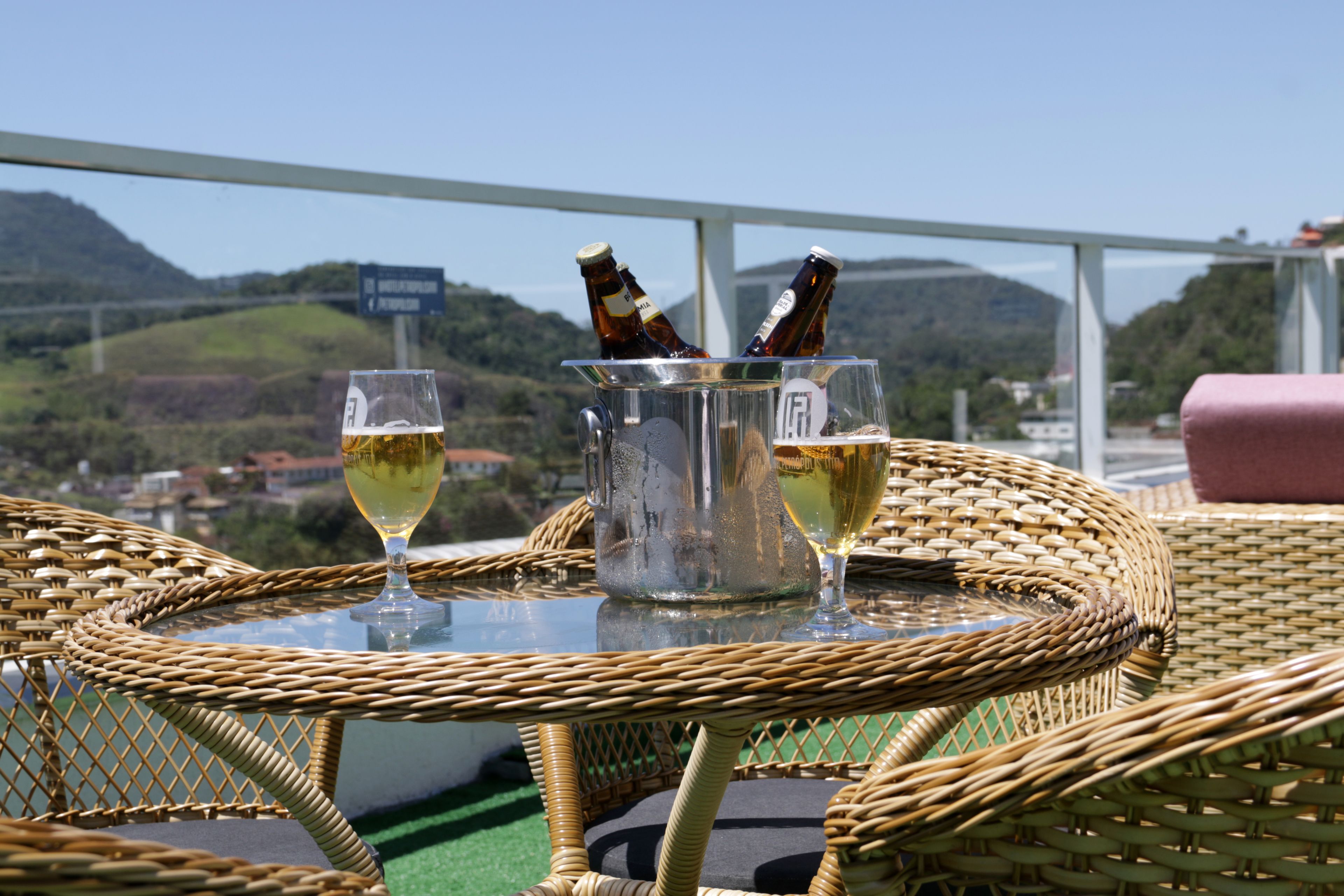 Una mesa de mimbre con dos copas de vino y una botella de cerveza, con vistas a colinas y un puente.