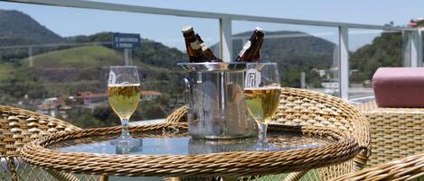 Una mesa de mimbre con dos copas de vino y una botella de cerveza, con vistas a colinas y un puente.