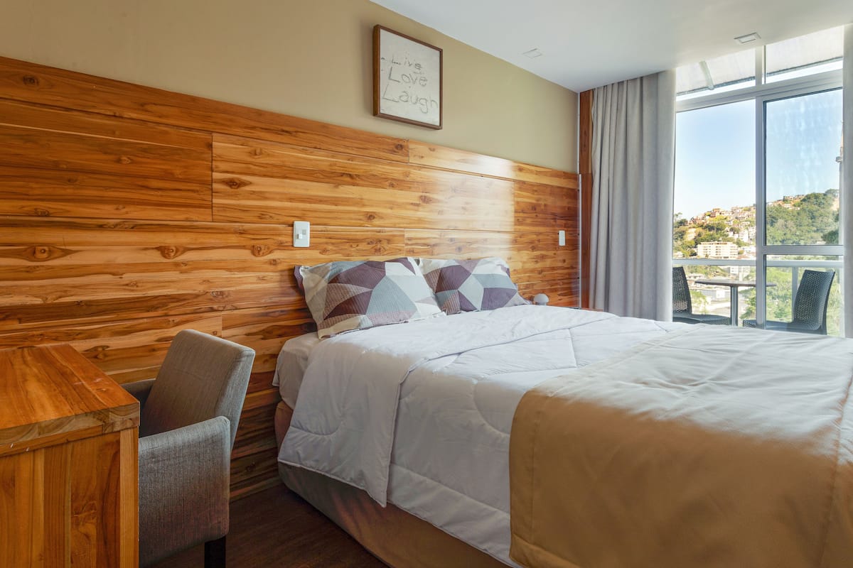 Habitación de hotel con cabecera de madera, cama con almohadas estampadas, un escritorio pequeño y vistas de un paisaje urbano a través de ventanas amplias.