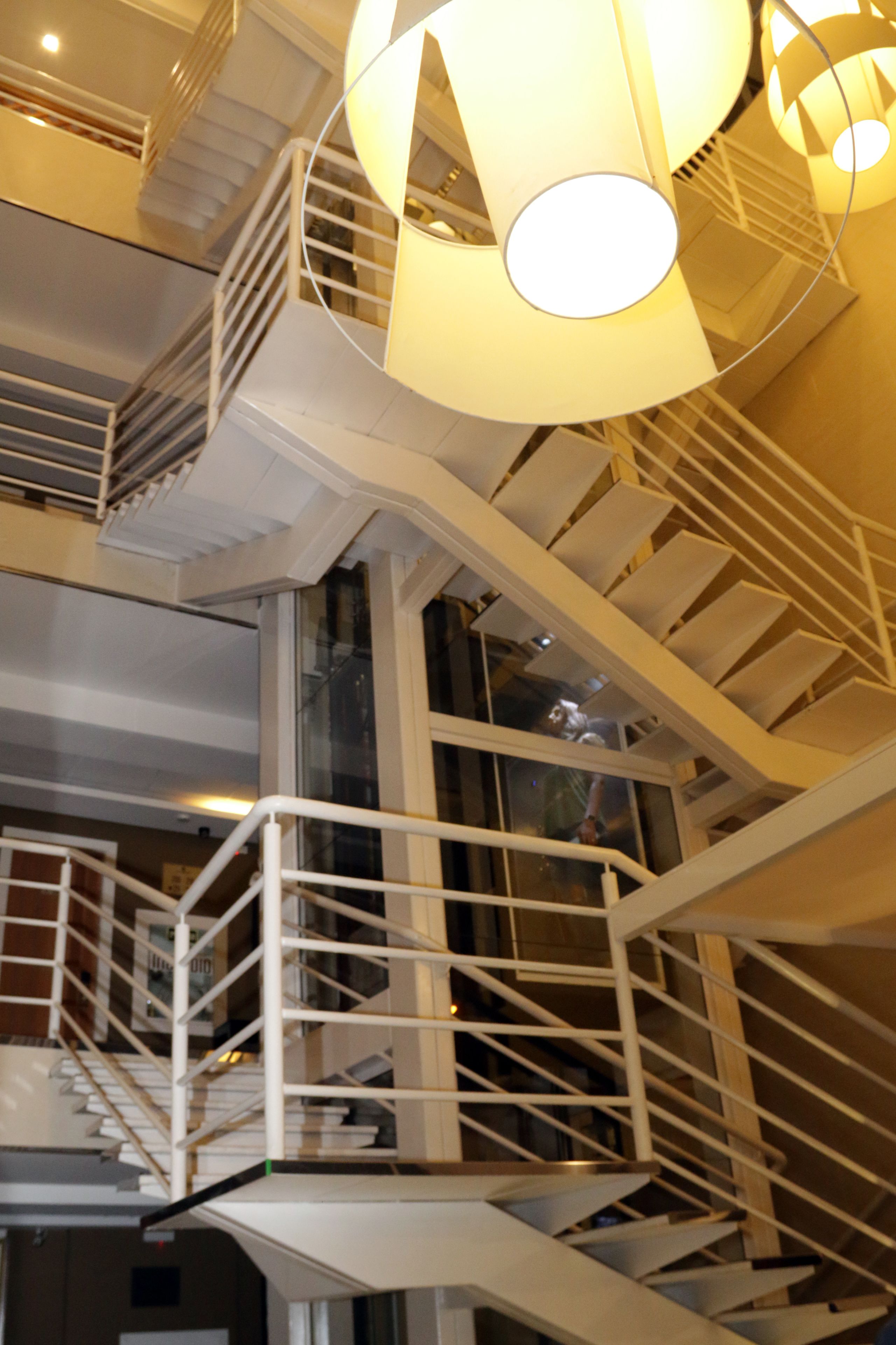 A multi-level interior with white staircases and railings, large circular light fixtures, and a glass elevator shaft.