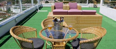 A rooftop terrace with wicker furniture, a glass table, and a bottle of beer.