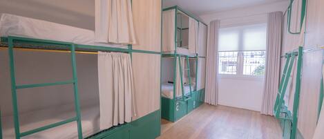 A dormitory-style room with bunk beds, a window with curtains, and a wooden floor.