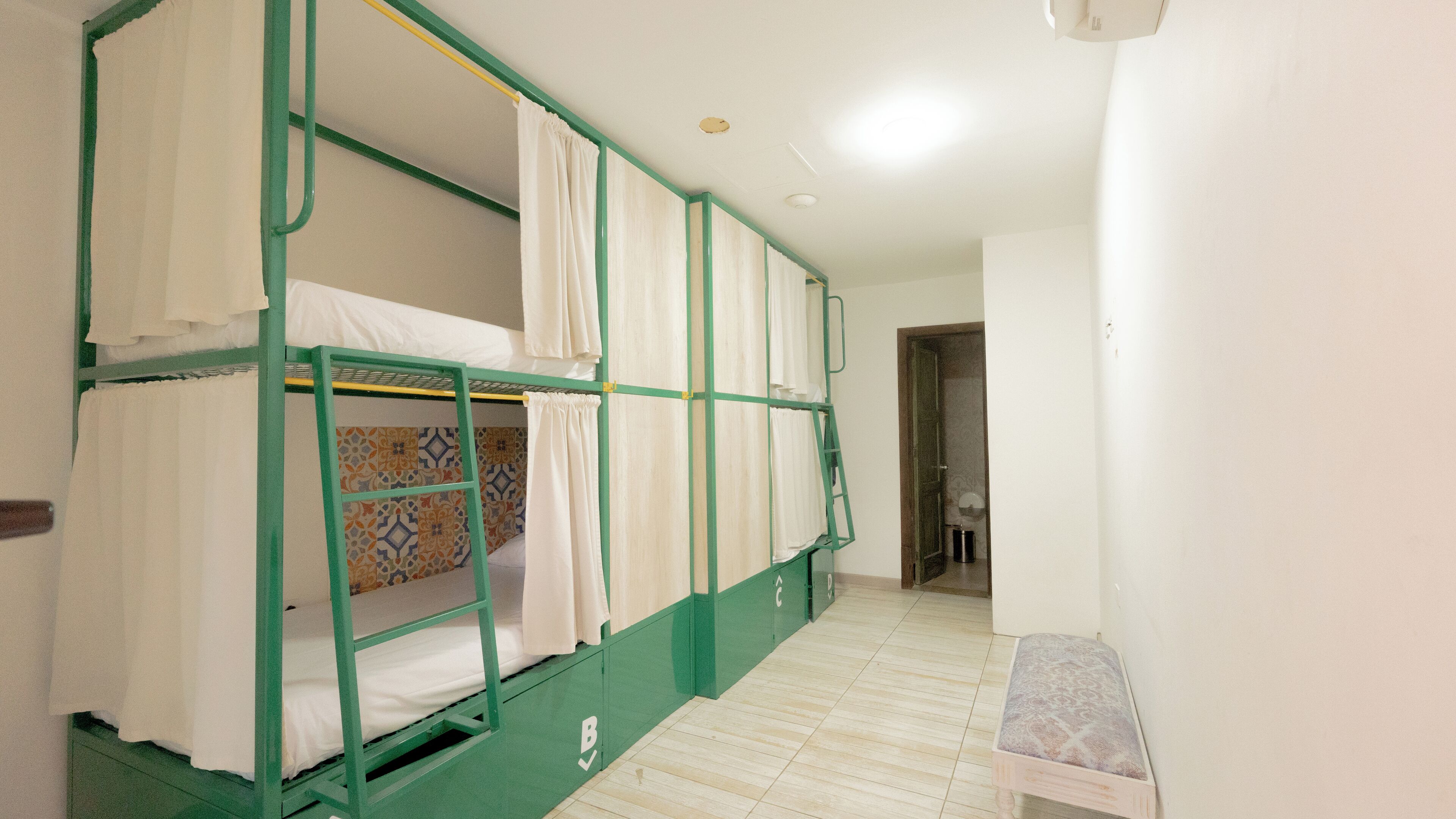 A dormitory room with bunk beds, a small bench, and a door leading to another room.