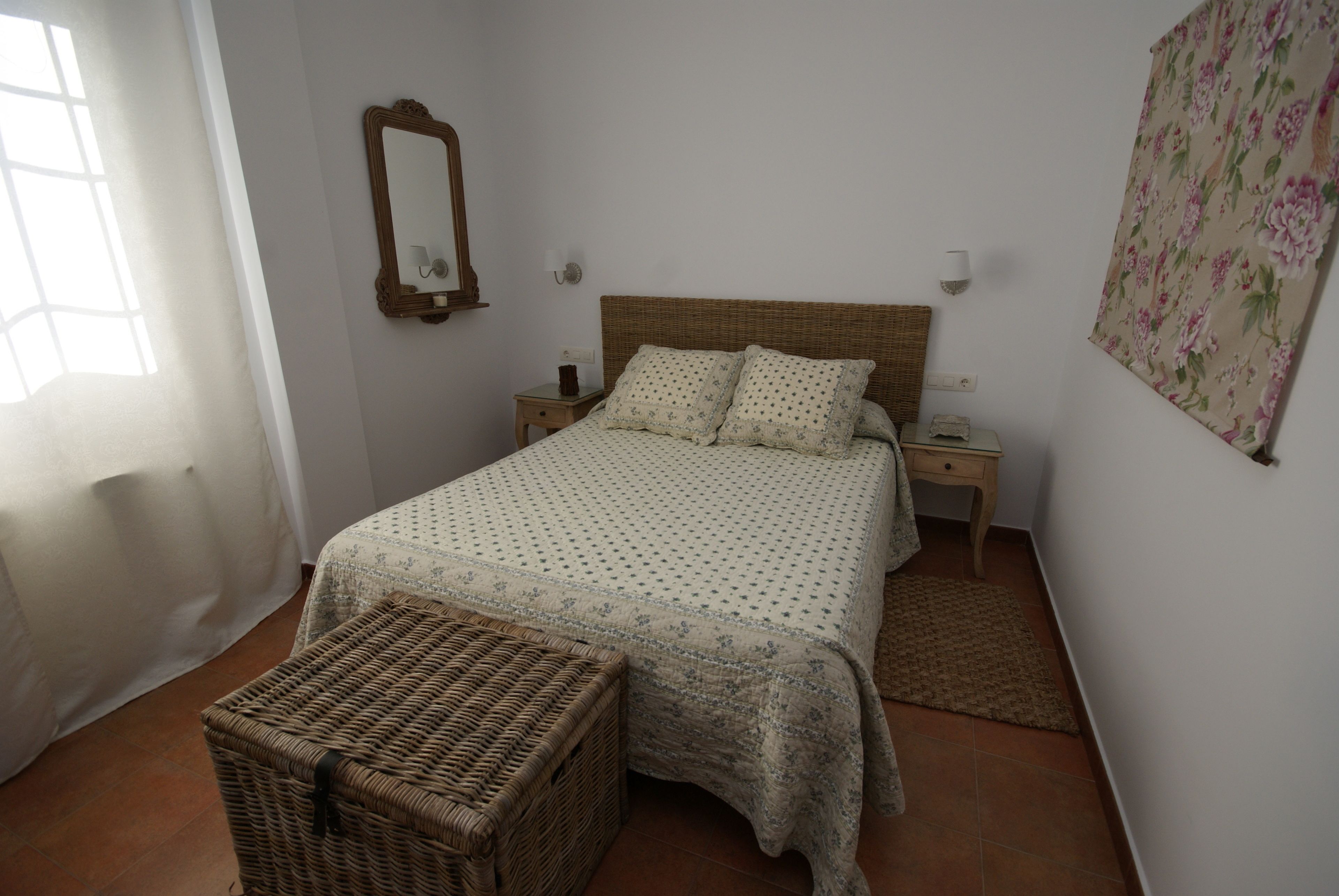 Un dormitorio con una cama, mesitas de noche, una cesta de mimbre, un espejo y un tapiz floral en la pared.