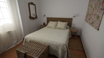 Un dormitorio con una cama, mesitas de noche, una cesta de mimbre, un espejo y un tapiz floral en la pared.