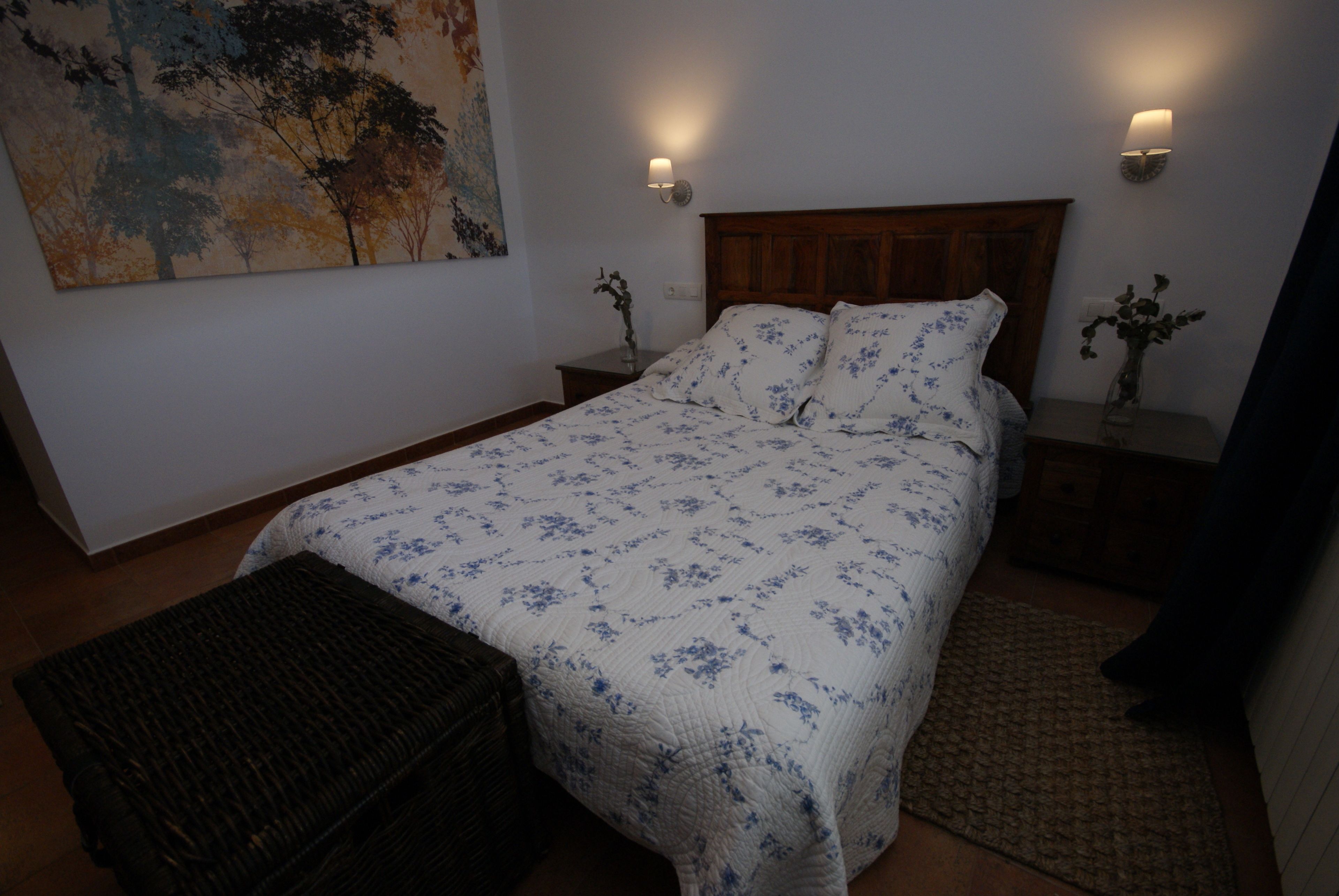 Una camera da letto con una testiera in legno, una coperta con motivi floreali, due lampade sui comodini, un comodino con un vaso e un quadro grande appeso al muro.