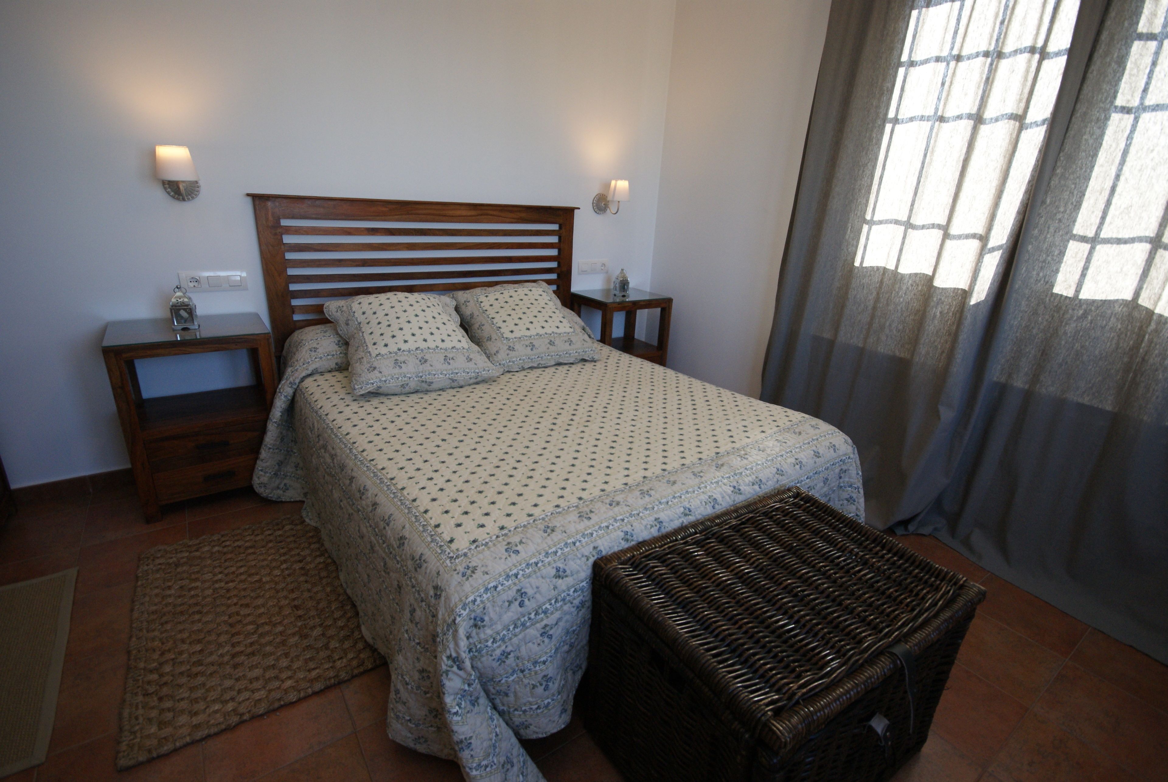A bedroom with a wooden headboard, a bed with a patterned bedspread, two bedside tables with lamps, a wicker ottoman, and a window with curtains.
