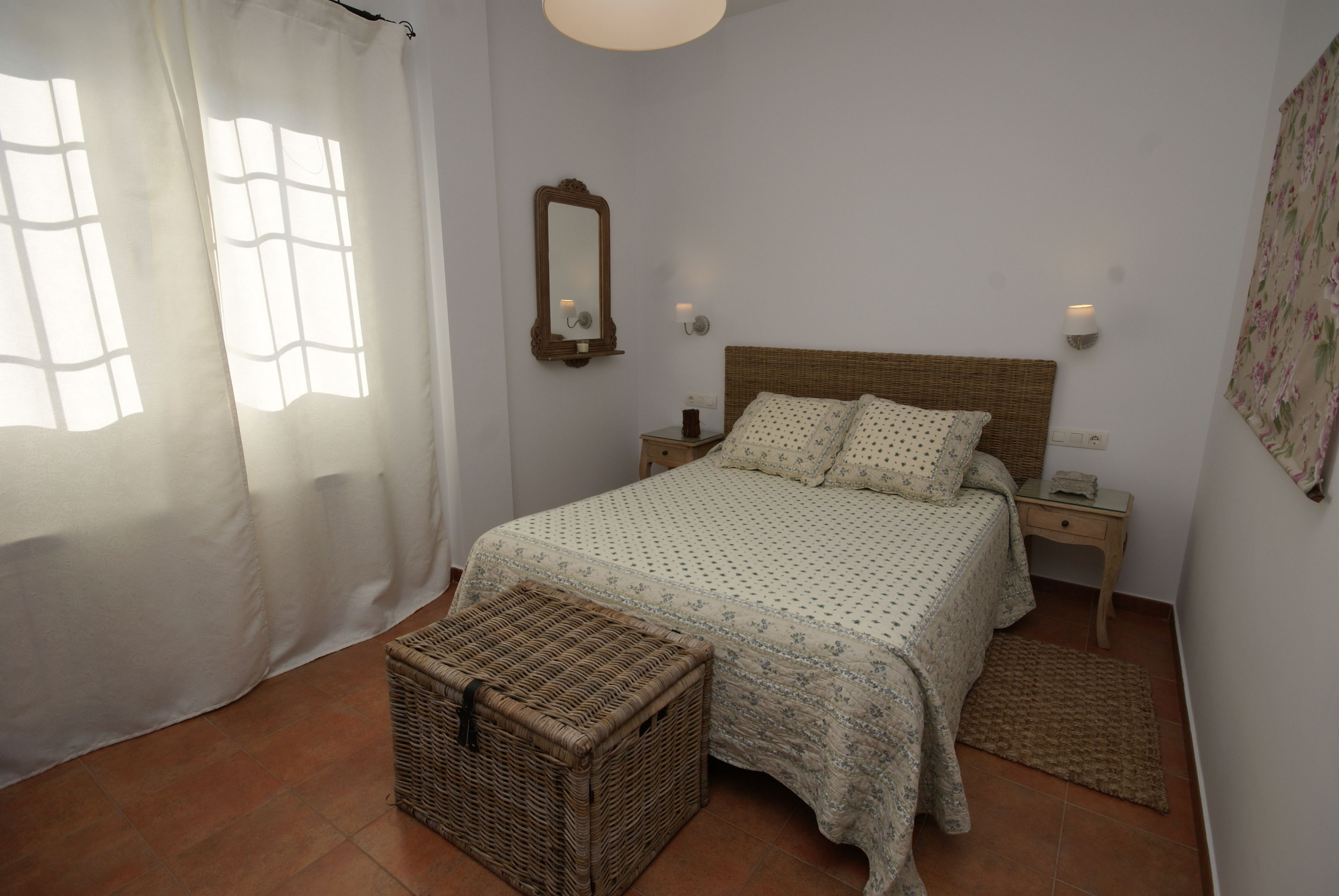 Un dormitorio con una cama, una mesita de noche, una cesta de mimbre, un espejo y una lámpara fijada a la pared.