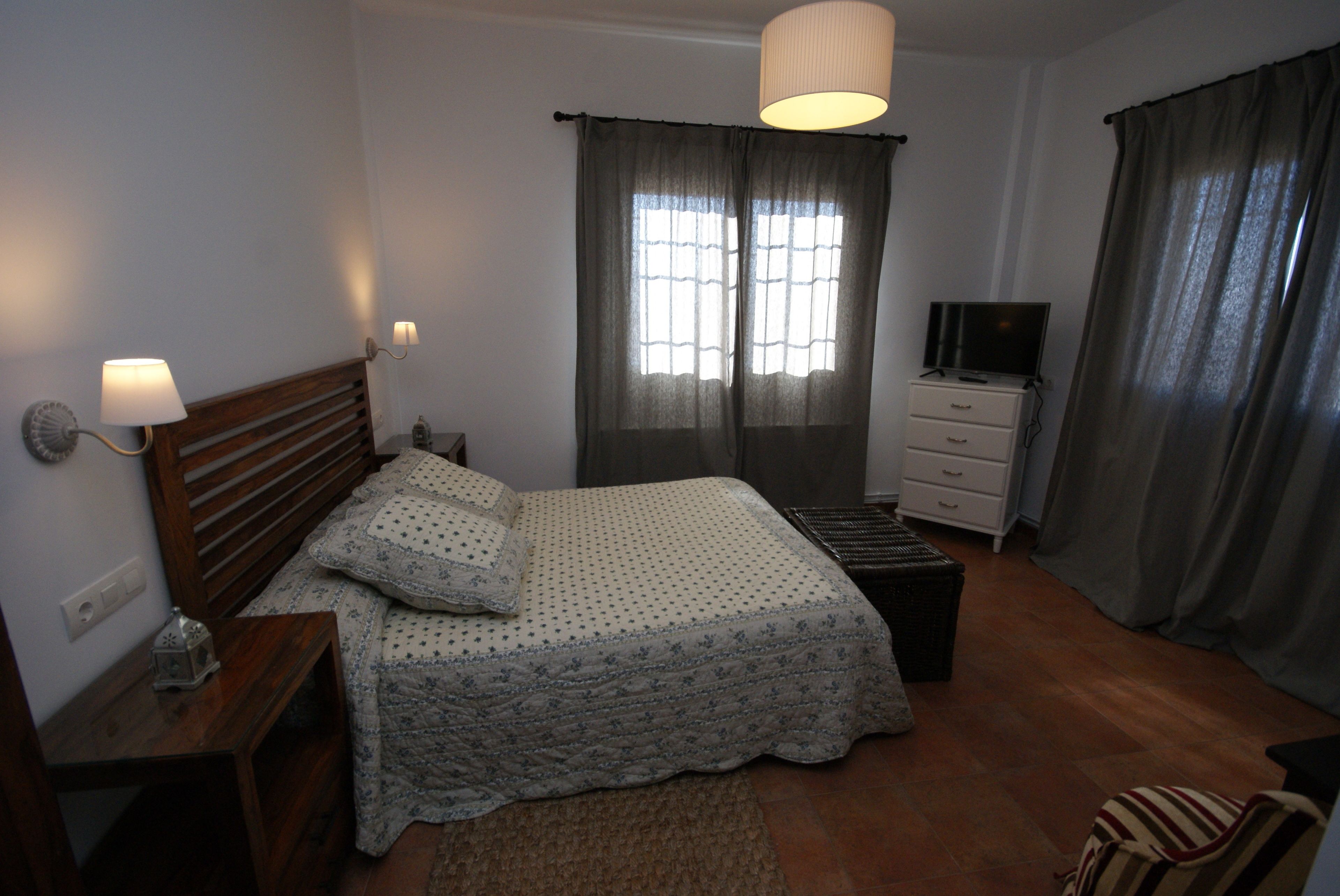 Un dormitorio con cama, mesita de noche, lámpara, televisor y cortinas.